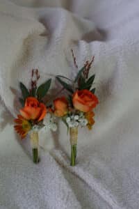 Zwei kleine Blumenboutonnieren mit orangefarbenen Rosen, weißem Schleierkraut und grünen Blättern sind zart umwickelte Stiele vor einem weichen, strukturierten cremefarbenen Stoffhintergrund – perfekt für jede Heiraten-Umgebung.