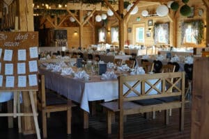 Ein rustikaler Bankettsaal, perfekt für eine Hochzeit, geschmückt mit Holzbalken und Dekorationen. Lange Tische mit weißen Tischdecken sind mit gefalteten Servietten und Blumendekorationen dekoriert. Ein Sitzplan wird auf einem Ständer angezeigt und sorgt für eine reibungslose Hochzeitsplanung.