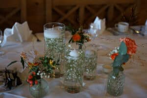 Ein Tischgesteck mit Glasvasen, in denen weiße schwimmende Kerzen und Schleierkrautblüten stehen, schafft eine perfekte Hochzeitsszene. Orange, rote und weiße Blüten mit Grün sorgen für Farbe. Im Hintergrund befinden sich gefaltete Servietten und Porzellantassen, ideal für jede Hochzeitsplanung.