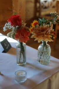Zwei Glasvasen mit orangefarbenen und roten Blumen, weißem Schleierkraut und Grünpflanzen stehen auf einem Hochzeitstisch. Im Vordergrund steht eine kleine, unbeleuchtete Kerze in einem Glashalter, während eine Tischnummernkarte mit der Aufschrift „51“ diesem Hochzeitstisch Charme verleiht.