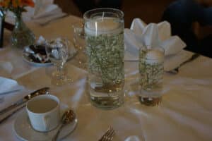 Die elegante Tischdekoration für die Hochzeit besteht aus zwei zylindrischen Glasvasen, die mit Wasser, schwimmenden Kerzen und zarten weißen Blumen gefüllt sind. Um die Vasen herum stehen eine weiße Teetasse mit Untertasse, poliertes Silberbesteck, ein Weinglas und eine kleine Vase voller leuchtend orangefarbener Blüten.