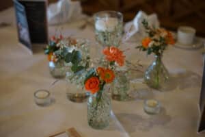 Ein Tischgedeck mit Glasvasen, in denen orangefarbene und weiße Blumen stehen, umgeben von kleinen Kerzen. Das Arrangement, perfekt für eine Hochzeit, steht auf einer weißen Tischdecke und schafft eine elegante und gemütliche Atmosphäre, ideal für Ihre Hochzeitsplanung.