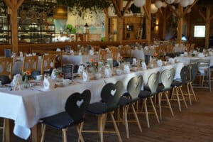 Ein rustikaler Bankettsaal mit langen Tischen, perfekt für Ihre Hochzeitsplanung. Jeder Tisch ist mit weißen Tischdecken, Blumendekorationen und ordentlich gefalteten Servietten geschmückt. Die Stühle haben herzförmige Rückenlehnen und der Raum verfügt über Holzbalken und sanfte Beleuchtung.