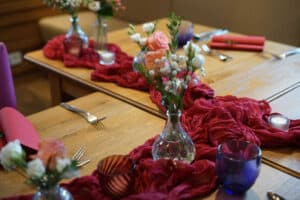 Ein hölzerner Esstisch ist elegant mit Vasen voller rosa und weißer Blumen dekoriert, perfekt für eine Hochzeitsfeier. Ein roter Stoffläufer und farbiges Glasgeschirr, darunter blaue und rote Gläser, verleihen eine lebendige Note. Silbernes Besteck und gefaltete rote Servietten vervollständigen dieses schöne Hochzeitsplanungsarrangement.