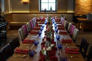 Ein langer Esstisch ist elegant für ein Hochzeitsfest gedeckt. Rote Servietten, blaue Gläser und Blumendekorationen schmücken den Tisch und schaffen eine festliche Atmosphäre, die perfekt für die Hochzeitsplanung ist. Der Raum ist warm beleuchtet, und natürliches Licht strömt durch ein Fenster am Ende des Raums.