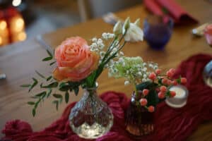 Ein Tisch, geschmückt mit zwei kleinen Vasen, eine mit einer rosa Rose und grünen Blättern, die andere mit weißen Blüten und roten Beeren. Ein roter Stoffläufer und ein kleines Teelicht werten das warme, elegante Ambiente auf – perfekt für alle Hochzeitsplanungsdetails.