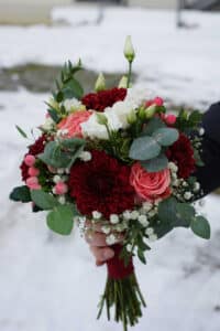 Eine Hand hält einen farbenfrohen Blumenstrauß vor einer verschneiten Kulisse, perfekt für die Hochzeitsplanung. Das Arrangement besteht aus roten und rosa Blumen, darunter Rosen und Dahlien, akzentuiert mit weißen Blüten und üppigen grünen Blättern und fängt die Essenz einer Traumhochzeit ein.