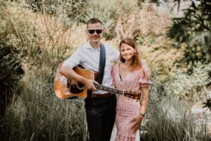 Ein Mann mit Sonnenbrille hält eine Gitarre und steht neben einer Frau im geblümten Kleid. Beide lächeln inmitten des üppigen Grüns von etwas, das wie ein Garten aussieht und vielleicht die Bühne für eine intime Hochzeitsfeier bereitet.
