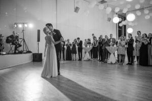 Ein Paar tanzt während eines Hochzeitsempfangs in der Mitte eines großen Raums. Im Hintergrund stehen die Gäste und schauen zu, einige applaudieren. Der Raum ist mit hängenden weißen Papierlaternen dekoriert, was exquisite Hochzeitsplanung widerspiegelt. Auf der Bühne links spielt ein Musiker Gitarre.