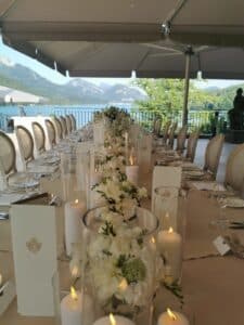 Ein langer, elegant gedeckter Tisch unter einem Baldachin ist mit weißen Blumendekorationen und brennenden Kerzen geschmückt. Mit Blick auf einen malerischen See und die Berge ist er perfekt für eine Hochzeit, mit ordentlich angeordneten Stühlen, die eine einladende Atmosphäre für ein Essen im Freien schaffen.