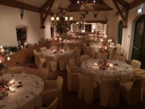 Der warm beleuchtete Bankettsaal, der sich perfekt für eine Traumhochzeit eignet, verfügt über runde Tische, die mit weißen Tischdecken und Blumendekorationen geschmückt sind. Elegante Kronleuchter und Kerzen bereichern das Ambiente, während rustikale Holzbalken und montierte Hirschgeweihe dieser idealen Kulisse für Hochzeiten eine charmante Note verleihen.