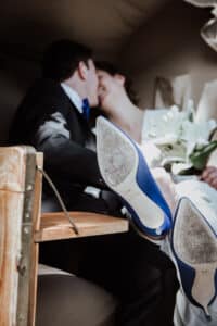 Ein Paar in formeller Kleidung teilt einen zärtlichen Moment, sitzend in einer Holzkutsche, die die Essenz einer Hochzeit verkörpert. Die Frau hält weiße Lilien und trägt blaue High Heels, die deutlich im Fokus stehen. Das Bild fängt die Intimität und Freude ihres bezaubernden Anlasses ein.