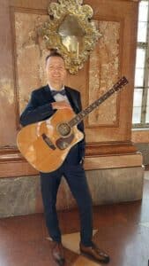 Ein lächelnder Mann im Anzug hält eine Akustikgitarre in der Hand. Er steht in einem Haus vor einer Marmorwand mit einem verzierten Spiegel über ihm. Durch ein Fenster rechts strömt natürliches Licht herein und er scheint bereit zu sein, bei einer Hochzeitsfeier ein Ständchen zu singen.