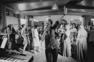 Auf einem Schwarzweißfoto ist eine Frau zu sehen, die in ein Mikrofon singt und mit einer Hand nach oben zeigt. Es sieht aus, als würde es sich um eine Hochzeitsfeier handeln. Sie befindet sich in einem überfüllten Raum, im Hintergrund tanzen fröhliche Gäste, während ein Keyboard zur lebhaften und festlichen Atmosphäre beiträgt.