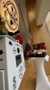 Ein Fotoautomat mit einer dekorierten Maschine, umgeben von Requisiten wie Hüten und Gläsern auf einem hohen, weißen Tisch. Weihnachtsdekorationen und ein Baum im Hintergrund deuten auf ein festliches Ereignis hin, das perfekt für jede Hochzeitsfeier ist.