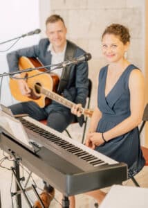 Eine Frau in einem marineblauen Kleid sitzt lächelnd an einer Tastatur und bereitet sich auf die Hochzeit vor. Vor ihr steht ein Mikrofon, während im Hintergrund ein Mann im Anzug Akustikgitarre spielt und lächelt. Sie befinden sich in einem Gebäude mit einer hellen Steinmauer im Hintergrund, die die perfekte Kulisse für ihren unvergesslichen Hochzeitsmoment bietet.