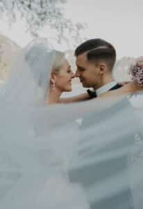 An ihrem Hochzeitstag umarmen sich Braut und Bräutigam im Freien. Die Braut trägt einen Schleier und hält einen Blumenstrauß, während der Bräutigam einen Anzug mit Fliege trägt. Vor der atemberaubenden Kulisse von Bäumen und Bergen blicken sie einander in die Augen und verlieren sich in der Magie von Heiraten.