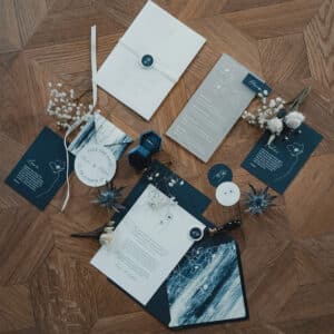 Eine flache Lage eleganter Hochzeitspapeterie auf einer Holzoberfläche, perfekt für jede Hochzeit, einschließlich Einladungen, Umschlägen und Karten mit Blumenillustrationen. Akzente sind ein Wachssiegel, kleine weiße Blumen und grüne Zweige.