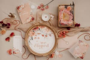 Eine flache Lage orangefarbener und rosa Hochzeitspapeterie auf beigem Stoff fängt die Essenz eleganter Hochzeitsplanung ein. Sie enthält ein „Wir sind durchgebrannt“-Schild, Einladungen, Umschläge, eine Kerze, Blütenblätter und dekorative Schnur, alles geschmückt mit floralen Mustern und eleganter Typografie.