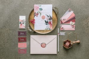 Eine flache Lage eleganter Hochzeitspapeterie auf einer grauen Oberfläche. Perfekt für Ihre Hochzeit. Es enthält Einladungen mit Blumenmotiven, einen Umschlag mit Wachssiegel, Namenskarten, eine sechseckige Schachtel und Farbmuster, die um ein Messingtablett angeordnet sind. Ideal, um Ihre exquisite Hochzeitsplanung zu präsentieren.