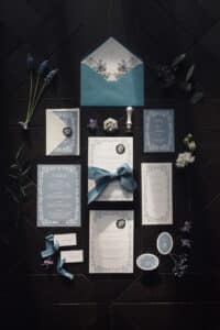 Eine Sammlung eleganter Hochzeitspapeterie mit Blumenmotiven in Blau und Weiß, perfekt für alle, die gerade mitten in der Hochzeitsplanung stecken, ist auf einer dunklen Holzoberfläche arrangiert. Das Set enthält Einladungen, Umschläge und kleine Dekorationsartikel, akzentuiert mit blauen Bändern und Grünpflanzen.