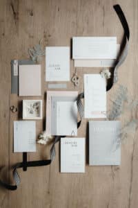 Eine Sammlung eleganter Hochzeitspapeterie, perfekt für Ihre Hochzeitsplanung. Präsentiert auf einer Holzoberfläche, enthält sie Einladungen, Menüs und RSVP-Karten in Pastelltönen mit minimalistischer Typografie. Verziert mit schwarz-weißem Band, Trockenblumen und zwei Ringen.