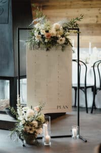Ein Hochzeitssitzplan auf einem Ständer, der oben mit weißen und rosa Blumen geschmückt ist, verkörpert Hochzeitsplanung perfekt. Kerzen und zusätzliche Blumenarrangements zieren den Sockel. Die Kulisse ist elegant, mit einer Holzkulisse und Esszimmerstühlen im Hintergrund, die die Romantik des Heiratens einfangen.