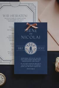 Elegante Hochzeitseinladungen für Ihre Hochzeit, bestehend aus einer blauen Karte mit „Irene und Nicolai“, einem dekorativen Emblem, dem 3. Dezember 2022 und einem Zitat von Astrid Lindgren. Darunter liegt eine teilweise sichtbare weiße Karte. Die Oberseite der blauen Karte ist mit einem Band geschmückt.