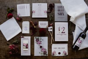 Eine Sammlung von Hochzeitsplanungs-Briefpapier mit Blumenmotiven auf einem Holztisch, darunter Einladungen, Menüs und Save-the-Date-Karten. Dekorative Elemente wie rote Blumen, eine Flasche Wein und Stoff tragen zur Ästhetik bei.