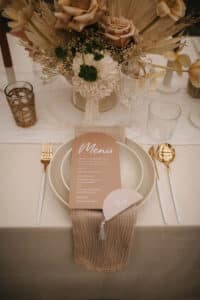 Ein wunderschön gedeckter Esstisch, perfekt für eine Hochzeit, ist in neutralen Tönen gehalten, mit einer Menükarte auf einem weißen Teller, gold-weißem Besteck und einer beigefarbenen Serviette. Getrocknete Blumenarrangements in sanften Farbtönen schmücken den Tisch und schaffen ein elegantes und rustikales Ambiente, ideal für die Hochzeitsplanung.