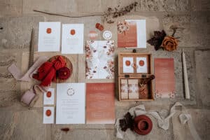 Flache Hochzeitspapeterie auf einer Steinoberfläche präsentiert elegante Hochzeitsplanung. Enthält Einladungen, Menüs und Save-the-Date-Karten in herbstlichen Farben, wunderschön geschmückt mit Bändern, Blumen, Wachssiegeln und einer großen weißen Kerze. Perfekte Details für Ihre Traumhochzeit.