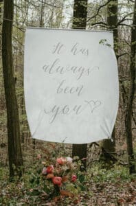 Ein weißes Banner hängt in einem Wald und zeigt den Text „Du warst es schon immer“ mit einem Herzsymbol. Darunter hängt ein Strauß bunter Blumen, der die Magie der Hochzeitsplanung in jedem Detail einfängt.