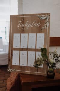 Ein hölzerner Sitzplan für Hochzeiten mit der Aufschrift „Tischplan“ an der Spitze verkörpert auf schöne Weise die Hochzeitsplanung und zeigt acht Tische mit der Aufschrift „Tisch 1“ bis „Tisch 8“. Auf jedem Tisch sind die Namen der Gäste aufgeführt. Mit zarten Blumendekorationen geschmückt, steht der Tisch elegant auf einem Ständer im Innenbereich.