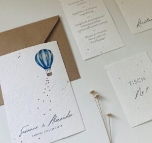 Hochzeitseinladungsset für Ihre Hochzeitsplanung bestehend aus einer Karte mit Heißluftballon-Motiv und den Namen „Susanne & Alexander“. Inklusive Umschlag, Menükarte und Tischnummernkarte, geschmückt mit zarten Trockenblumen auf weißem Untergrund. Termin: Samstag, 3. Juli 2023.