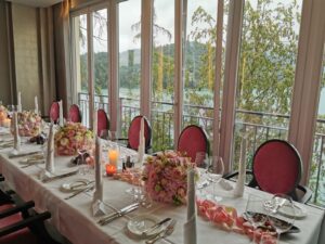 Ein wunderschön gedeckter Esstisch mit weißen Tischdecken, rosa Blumenarrangements und Kerzen sorgt für eine perfekte Hochzeitsatmosphäre. Durch die großen Fenster mit Blick auf üppiges Grün fängt er die Romantik von Heiraten mit Blick auf Bäume und ferne Hügel ein.