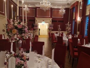 Ein eleganter Bankettsaal, perfekt für Ihre Hochzeit, mit roten Stühlen und weißen Tischdecken. Die Tische sind mit großen Blumenarrangements aus rosa und weißen Blumen geschmückt. Kristallkronleuchter erhellen den Raum, während die Wände in sattem Rot mit weißen Verzierungen eine atemberaubende Kulisse für unvergessliche Momente schaffen.