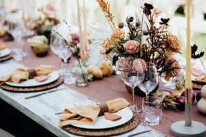 Eine rustikale Tischdekoration im Freien, perfekt für eine Hochzeit, mit Tischsets aus Korbgeflecht, gefalteten Servietten, Glaswaren und Blumengestecken. Geschmückt mit Kerzen und einer Auswahl an herbstlichen Blumen und Blättern bietet das Ambiente einen gemütlichen und festlichen Charme, ideal für die Hochzeitsplanung.