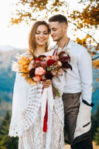 Ein Paar steht eng beieinander im Freien und verkörpert seine Hochzeit. Die Frau, die einen Strauß bunter Blumen hält und ein Spitzenkleid trägt, steht im Mittelpunkt des liebevollen Blicks ihres Partners. Sonnenlicht fällt durch die Herbstblätter im Hintergrund und fängt die Essenz zeitloser Liebe ein.