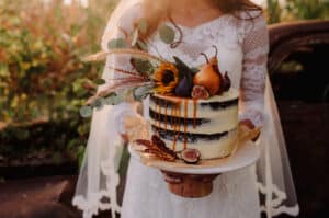 Eine Frau in einem weißen Spitzenkleid hält einen rustikalen, teilweise mit Zuckerguss überzogenen Kuchen auf einem Holzständer. Der Kuchen ist perfekt für eine Hochzeit und mit Karamell, Feigen, Birnen und Herbstlaub verziert. Sonnenlicht beleuchtet die Szene und schafft eine warme, natürliche Atmosphäre, die ideal für Momente der Hochzeitsplanung ist.