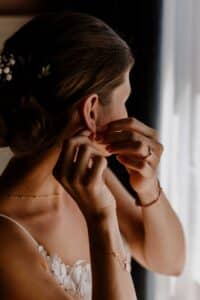 Eine Frau mit hochgestecktem Haar setzt Ohrringe auf und bereitet sich auf eine wunderschöne Hochzeit vor. Sie trägt eine zarte Halskette und ein mit Spitze besetztes Outfit und steht neben einem Fenster, wo sanftes Licht sie sanft beleuchtet und die Eleganz sorgfältiger Hochzeitsplanung verkörpert.