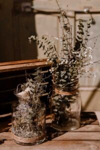 Zwei mit getrockneten Eukalyptuszweigen und Schleierkraut gefüllte Glasgefäße stehen auf einer Holzfläche – perfekt für eine rustikale Hochzeit. Die Gläser sind mit Schnur umwickelt und die Szene ist in warmes, natürliches Licht getaucht, das weiche Schatten wirft.