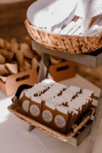 Ein Weidenkorb auf einem Holzständer hält Papierfächer über einem weiteren Korb voller kleiner brauner Tüten mit der Aufschrift „Freudentränen“, perfekt für Ihre Hochzeit. Im unscharfen Hintergrund warten weitere Tüten als Teil einer durchdachten Hochzeitsplanung.