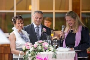 Ein Paar steht während seiner Hochzeit an einem wunderschön dekorierten Tisch. Die Braut hält einen Blumenstrauß und der Bräutigam steht neben ihr. Beide lächeln strahlend. Eine Frau mit einem Mikrofon und einem rosa Schal spricht fröhliche Worte. Im unscharfen Hintergrund sind Hinweise auf eine sorgfältige Hochzeitsplanung zu erkennen.