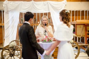 Eine romantische Hochzeit findet in einem rustikalen Rahmen statt. Ein Paar hält sich an den Händen und blickt dem Geistlichen gegenüber. Der Geistliche steht vor üppigen Blumenarrangements und spricht in ein Mikrofon. Vintage-Wagenräder und ein weißer, drapierter Hintergrund bilden die perfekte Kulisse für diese wunderschön geplante Hochzeit.