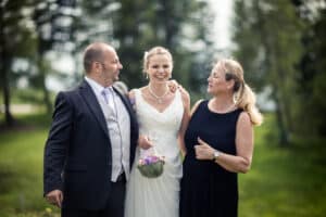 Inmitten des üppigen Grüns steht eine Braut in einem weißen Kleid zwischen einem Mann im Anzug und einer Frau in einem dunklen Kleid. Mit strahlenden Lächeln umarmen sie sich und feiern die Freude des Heiratens. Die Braut hält einen kleinen Blumenstrauß in der Hand, der die Essenz dieses wunderschön geplanten Hochzeitsmoments im Freien einfängt.
