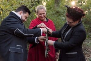 Drei Personen nehmen im Freien an einem zeremoniellen Handfasting-Ritual teil, das einer bezaubernden Hochzeit ähnelt. Der Mann und die Frau halten Händchen und lächeln, während eine Frau in einem roten Umhang hilft, Bänder um ihre Hände zu binden. Die Kulisse ist ein üppiger, sonnenbeschienener Wald, perfekt für eine einzigartige Hochzeitsplanung.