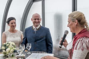 Ein Paar steht während einer eleganten Hochzeitszeremonie einer Frau gegenüber, die ein Mikrofon hält. Die Braut trägt ein Spitzenkleid und hält einen Blumenstrauß, während der Bräutigam einen traditionellen Anzug trägt. Sie sind drinnen, mit großen Fenstern im Hintergrund, und fangen diesen besonderen Moment ihrer Heiraten-Reise ein.