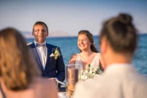 Braut und Bräutigam lächeln freudig bei einer Hochzeitszeremonie im Freien am Strand. In formeller Kleidung feiern sie ihre Hochzeit mit dem Meer im Hintergrund, was die sorgfältige Hochzeitsplanung lohnenswert macht. Andere Gäste sind teilweise im Vordergrund zu sehen.