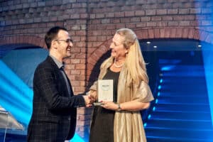 Ein Mann und eine Frau lächeln und schütteln sich bei einer Hochzeitsfeier die Hände. Die Frau hält eine Plakette in der Hand, die möglicherweise mit ihrer außergewöhnlichen Hochzeitsplanung zusammenhängt. Sie stehen vor einer blau beleuchteten Backsteinmauer und wirken beide zufrieden und feierlich.