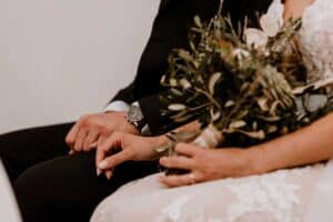 Braut und Bräutigam sitzen während ihrer wunderschönen Hochzeit zusammen. Die Braut hält einen Strauß Grünpflanzen und Blumen, während der Bräutigam eine Uhr trägt. Beide legen ihre Hände sanft aneinander und fangen einen ruhigen Moment ihrer Hochzeitsreise ein.
